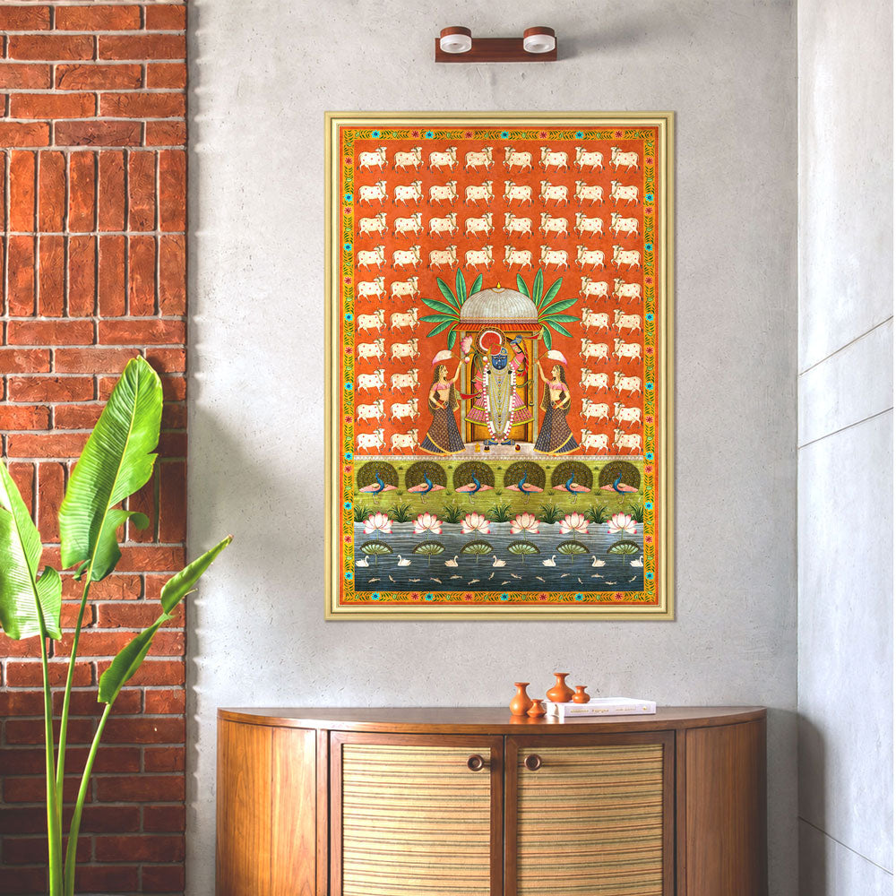 A framed colorful wall art print depicting a traditional Hindu deity scene, displayed on a light gray wall above a wooden cabinet in a styled interior setting with a brick wall and plant.