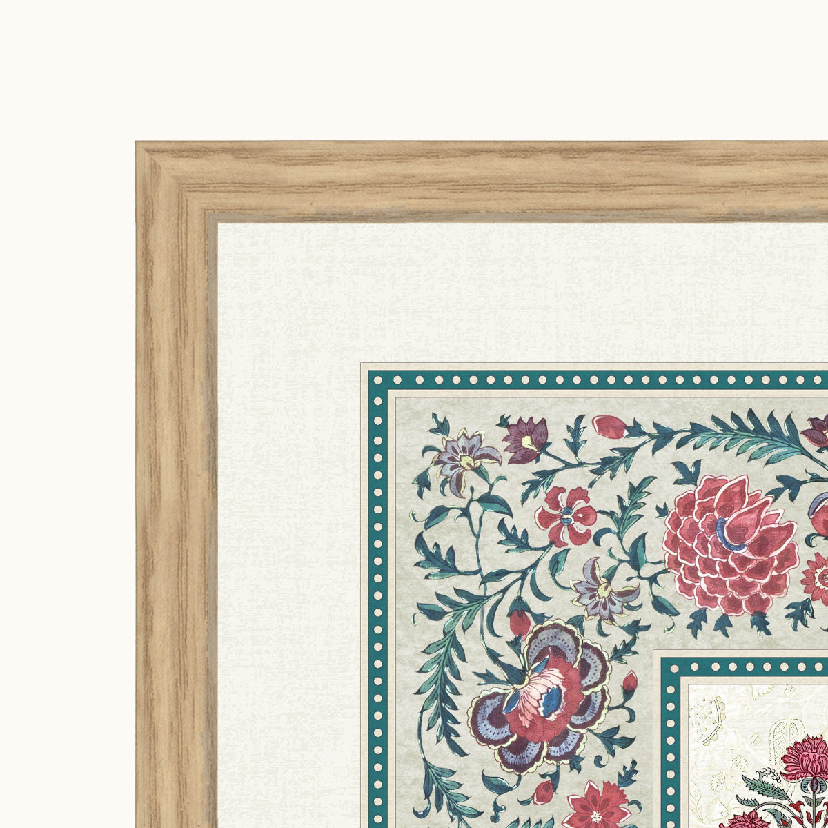 Close-up of a framed floral artwork showing part of a light wood frame, white mat, and a cream rug or textile with teal, pink, and blue botanical motifs.