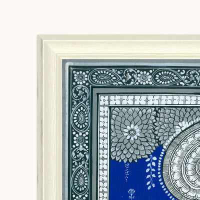 A close-up of a framed decorative art showing the corner of the frame and part of the artwork. The print features intricate floral and geometric patterns in gray, white, and dark green on a vivid blue background.