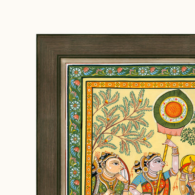 Close-up of a framed Indian folk art showing painted figures, a tree, and decorative borders, with a dark wood frame on a light background.